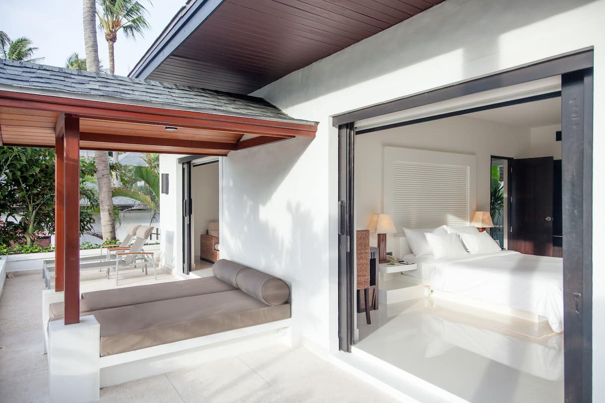 A modern bedroom with a large bed, a seating area with a sofa and chairs, and a glass door leading to a balcony.