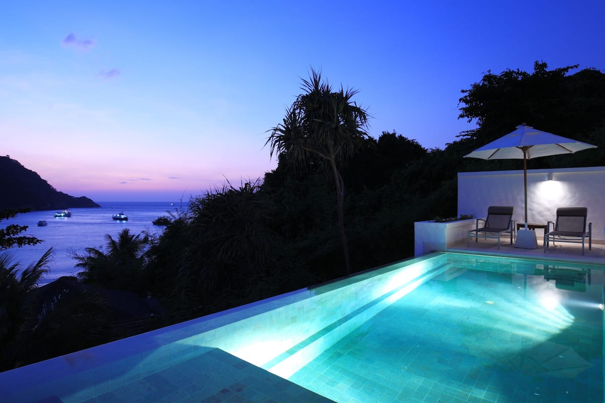 A swimming pool with a view of the ocean at dusk, surrounded by palm trees and a lounge area with chairs and an umbrella.