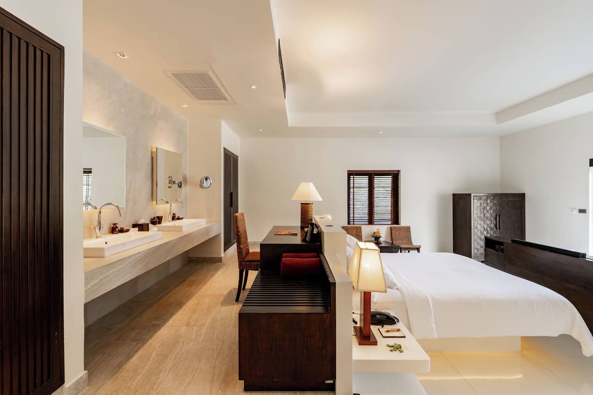 A modern hotel room with a large bed, a desk with a lamp, a chair, a TV, and a bathroom with two sinks and a mirror.