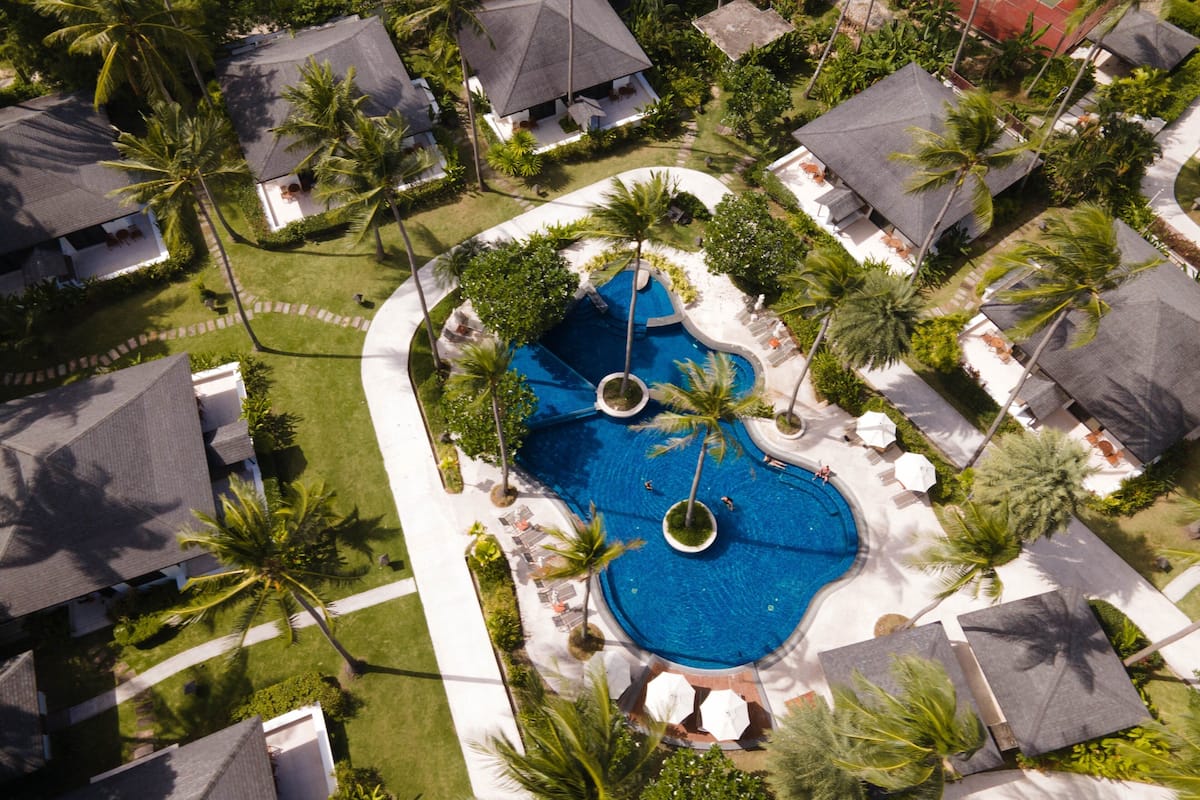 An aerial view of a resort with a central swimming pool, surrounded by palm trees and covered outdoor seating areas.