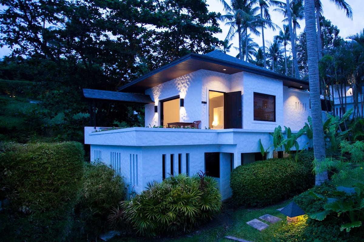 A modern house with a balcony, surrounded by lush greenery and palm trees.