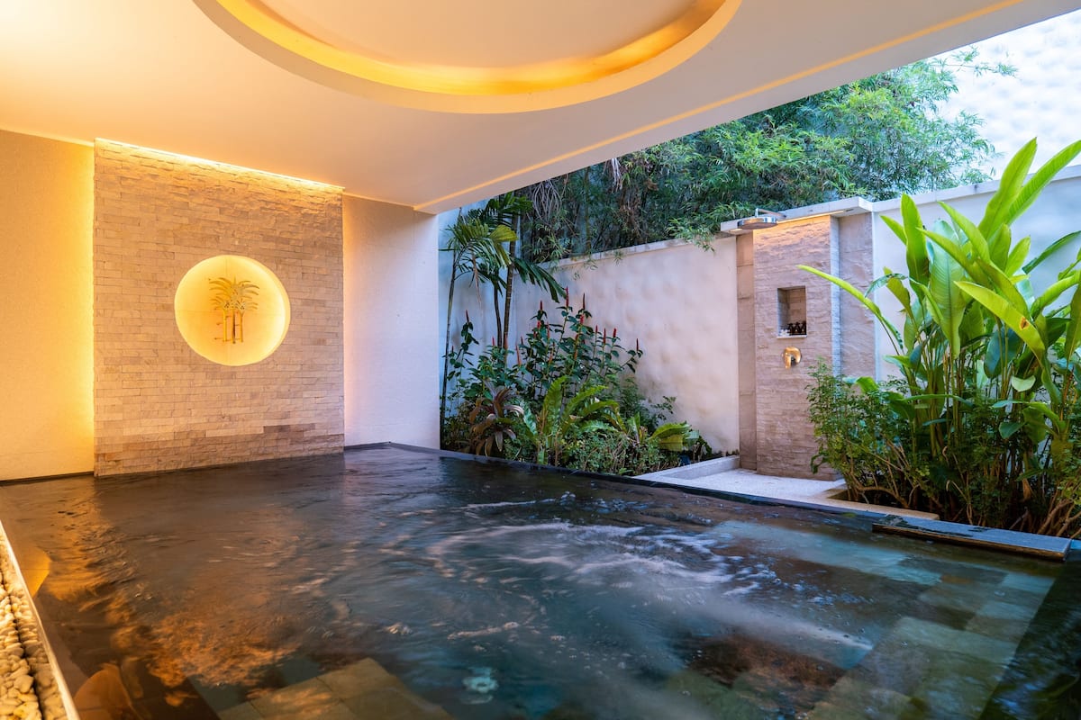 A spa area with a hot tub, a brick wall with a circular light fixture, and lush greenery.