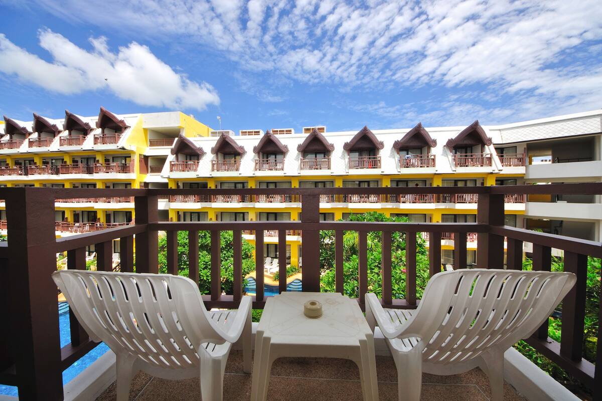 A hotel balcony with white plastic chairs and a table, overlooking a pool and lush greenery.