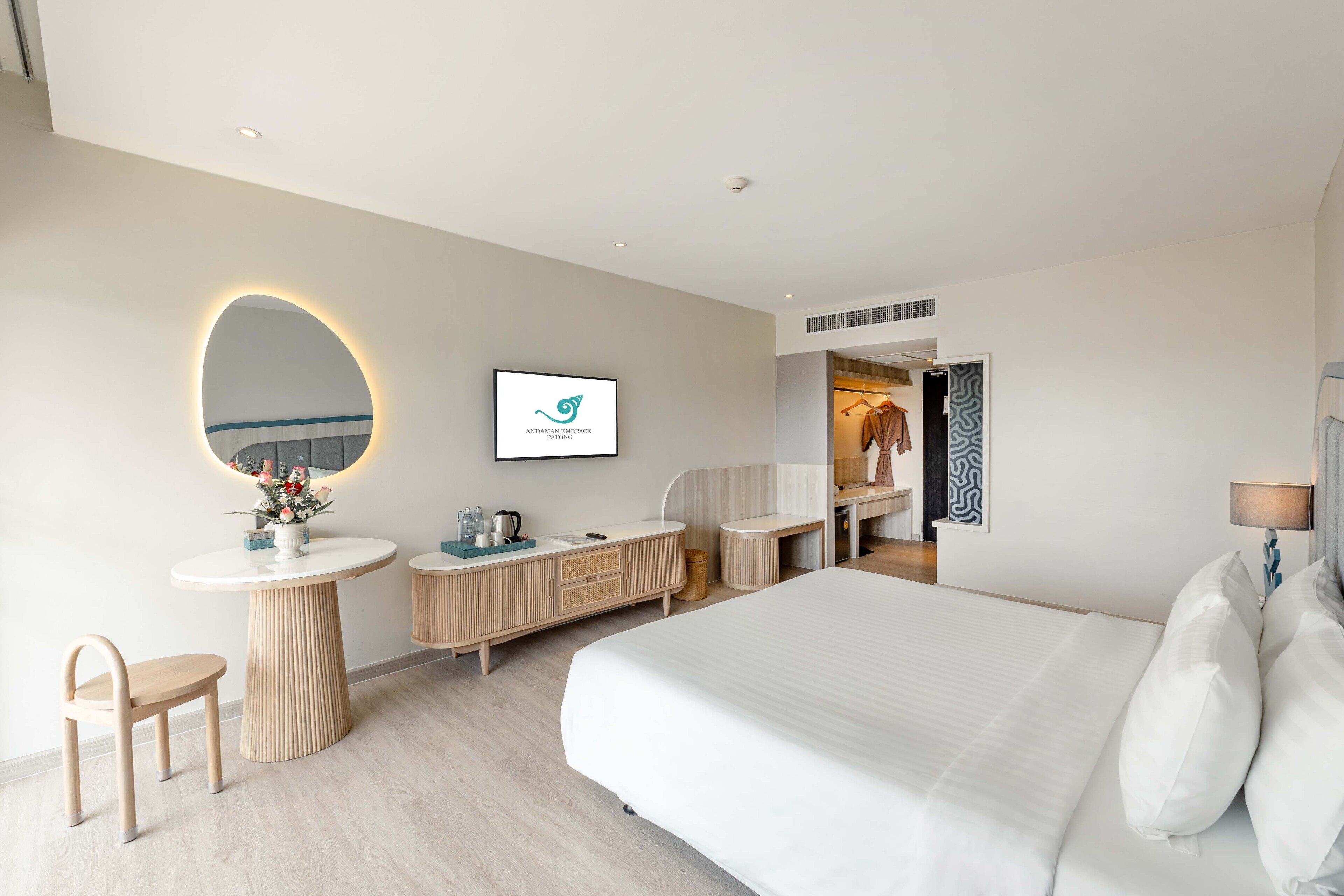 A modern hotel room with a large bed, a round mirror, a vanity with a sink, a wooden cabinet, and a small table with a vase of flowers.