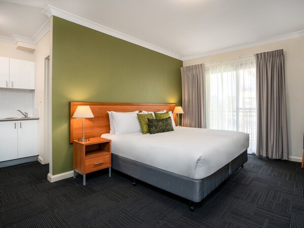 Une chambre d’hôtel avec un grand lit, une table de chevet en bois avec une lampe, un petit bureau avec une chaise, une kitchenette et une fenêtre avec des rideaux.