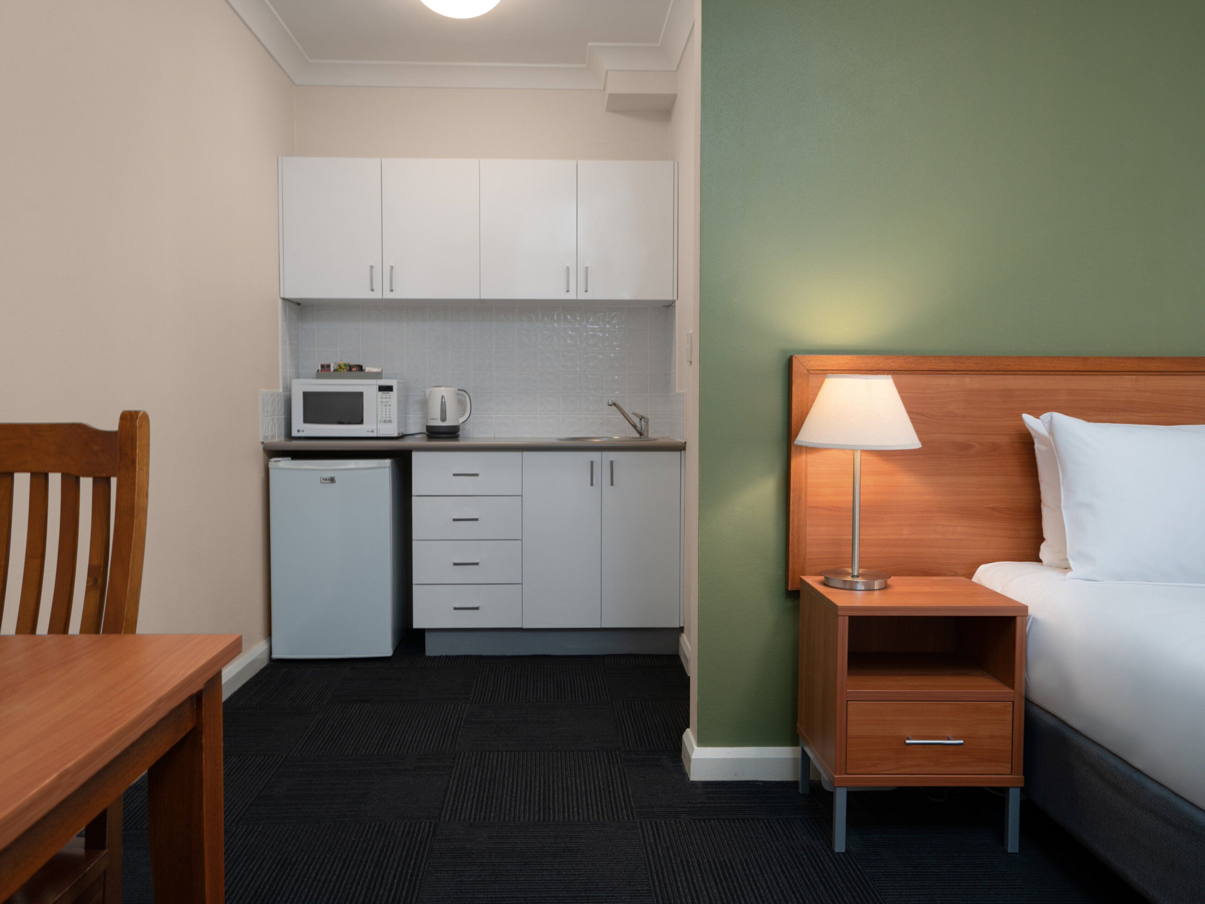 A compact hotel room with a kitchenette, a bed, a nightstand, and a wooden chair.