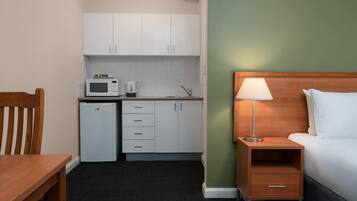 Una habitación de hotel compacta con kitchenette, una cama, una mesita de noche con lámpara y una silla de madera.