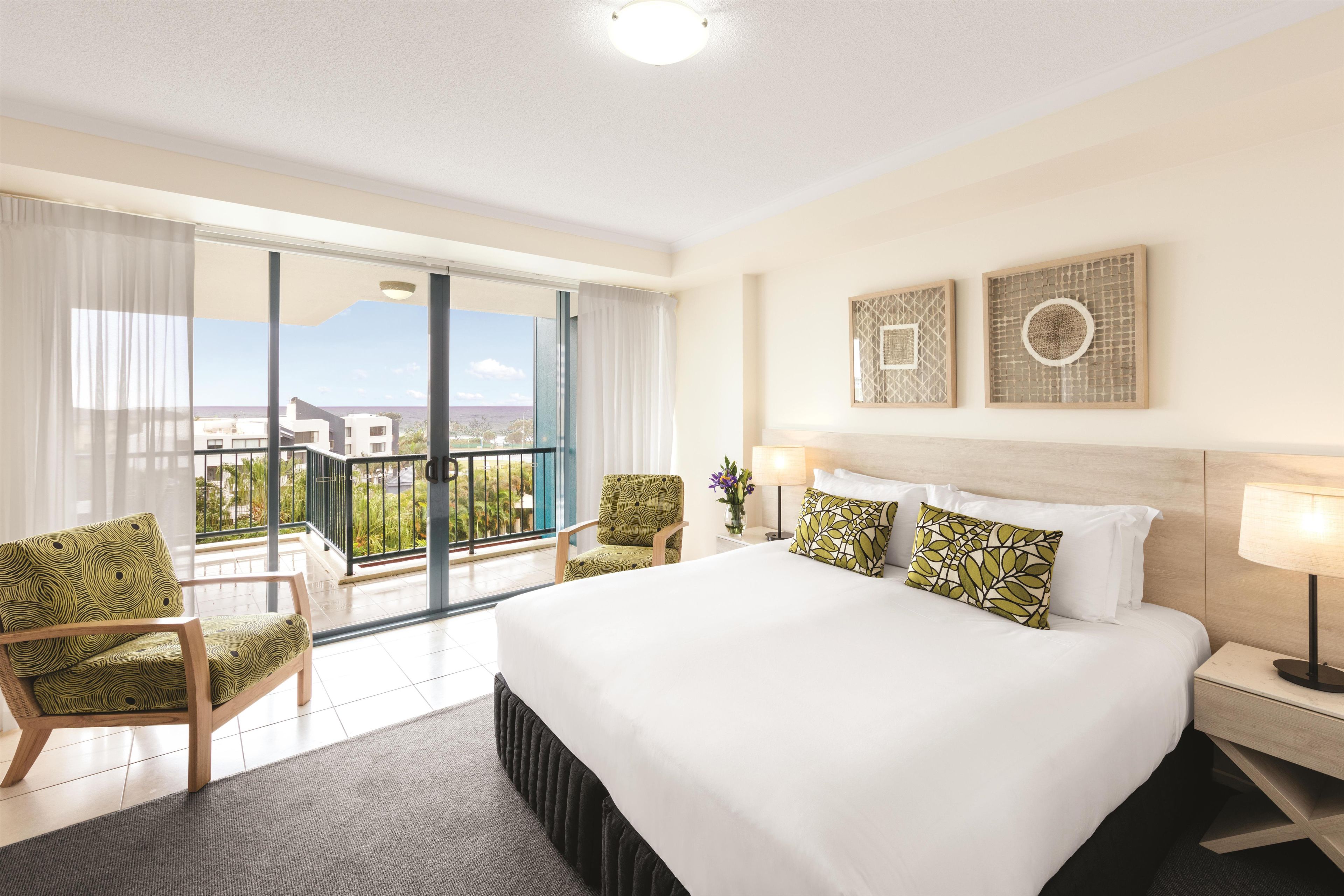 Habitación de hotel con una cama grande, una zona de estar con sillas, un balcón con vistas y dos obras de arte colgadas en la pared.