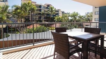 Een balkon met een tafel en stoelen, met uitzicht op een zwembadgedeelte met palmbomen en gebouwen op de achtergrond.