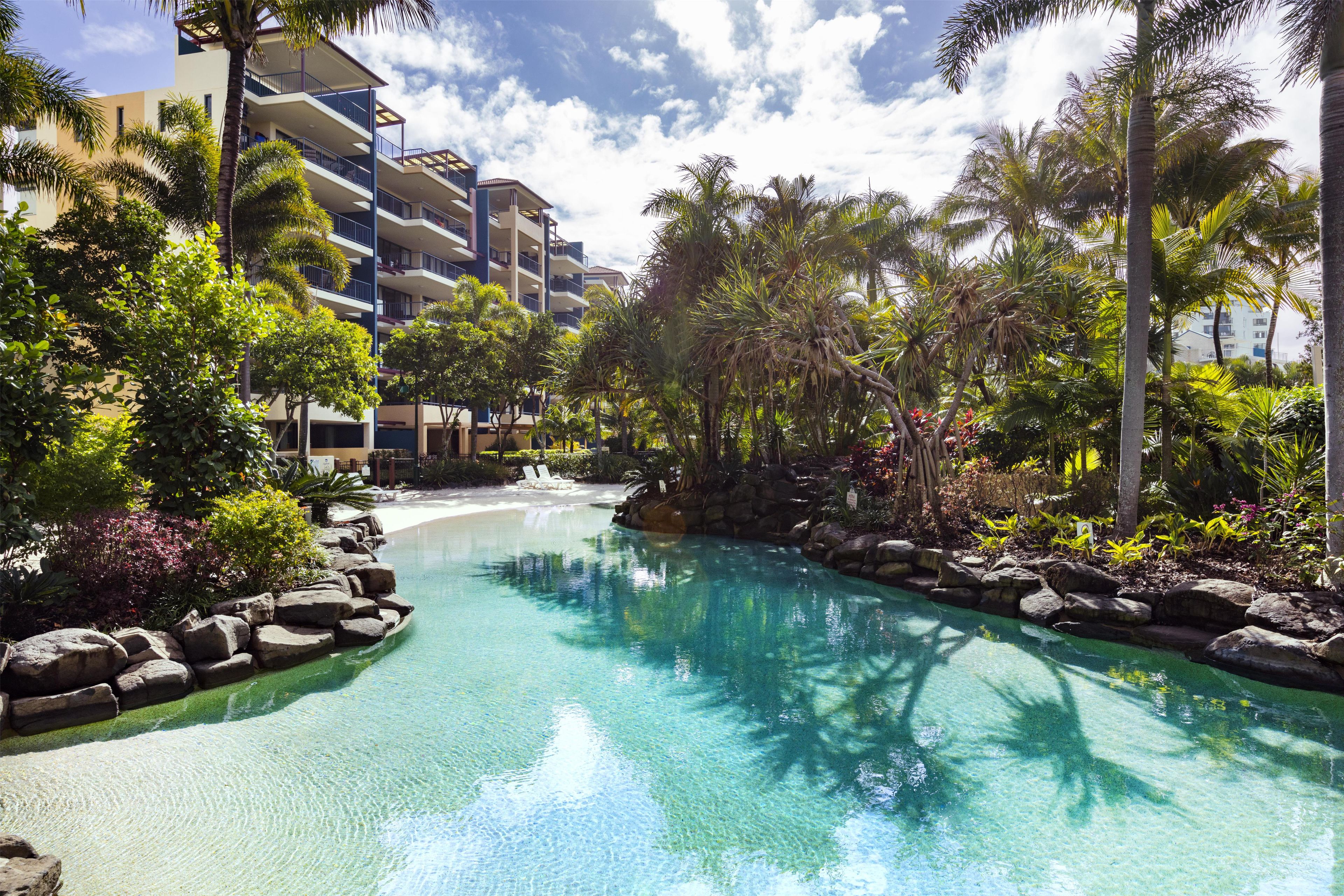 A resort pool with clear water, surrounded by tropical vegetation and a multi-story building.
