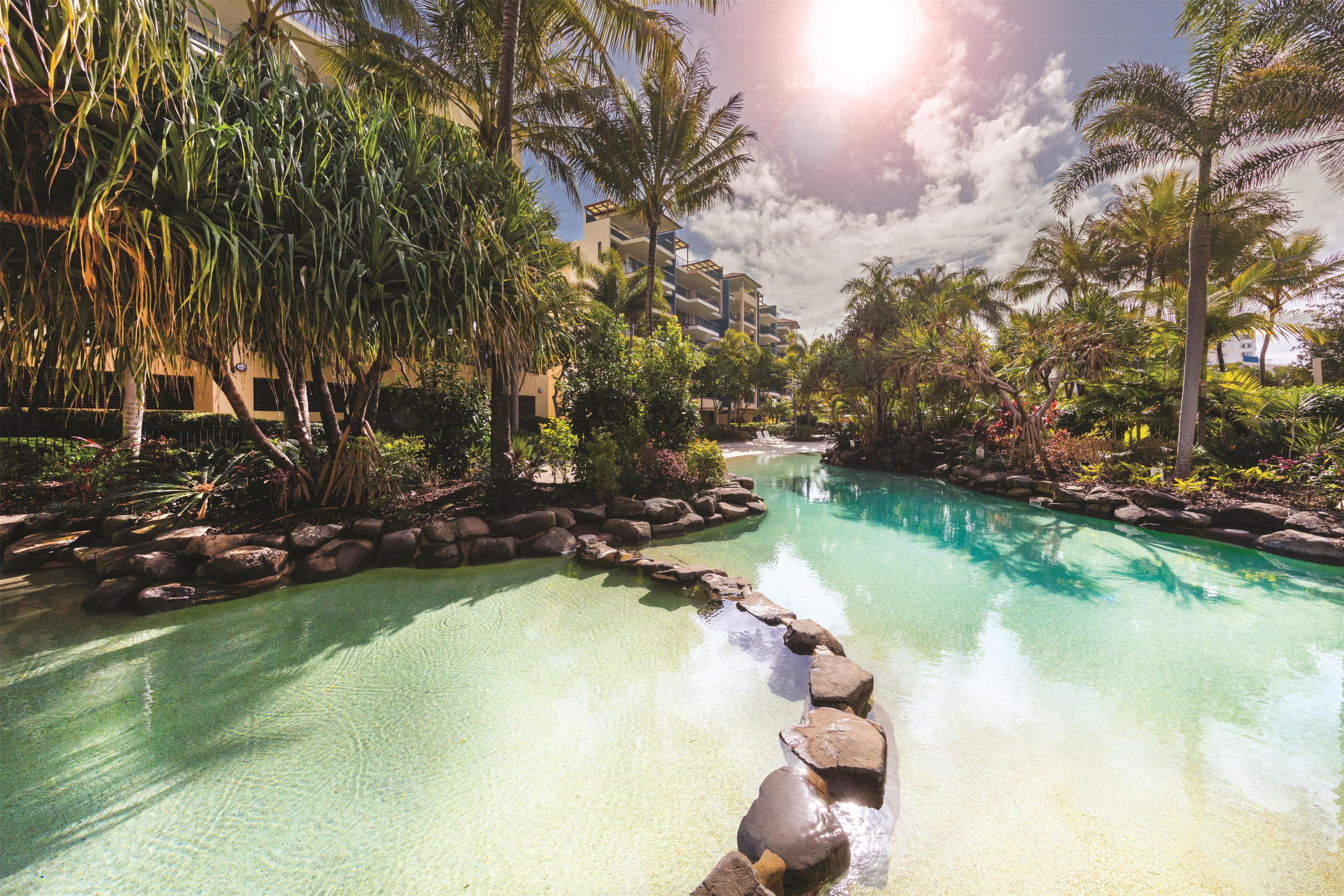 A clear pool with rocks in the foreground, surrounded by lush greenery and palm trees, with a building in the background.