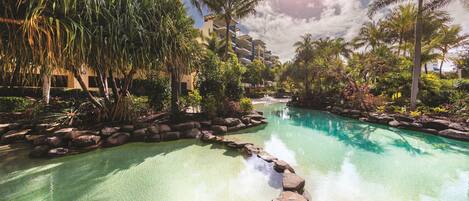 A clear pool with rocks in the foreground, surrounded by lush greenery and palm trees, with a building in the background.