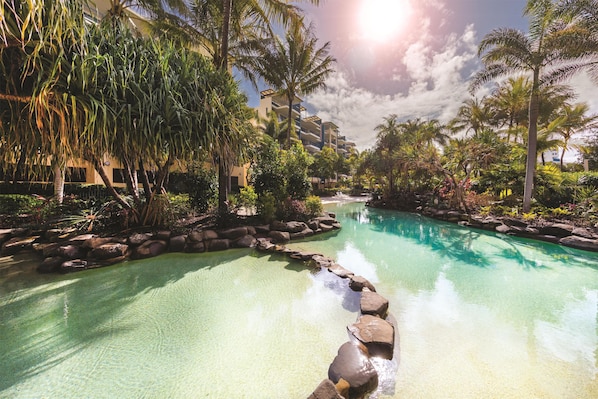 Una piscina cristalina con rocas al frente, rodeada de exuberante vegetación y palmeras, con un edificio en el fondo.