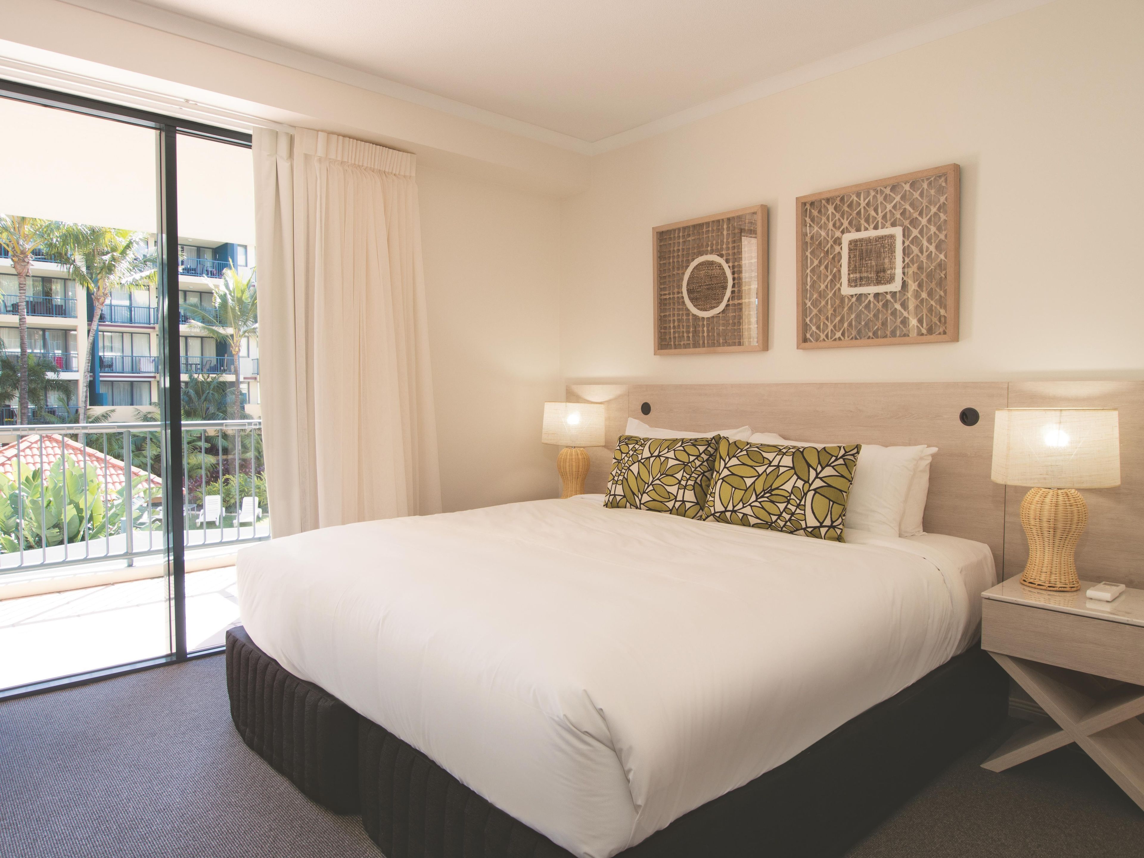 A hotel room with a large bed, two bedside lamps, a view of a building with balconies, and two framed wall art pieces.