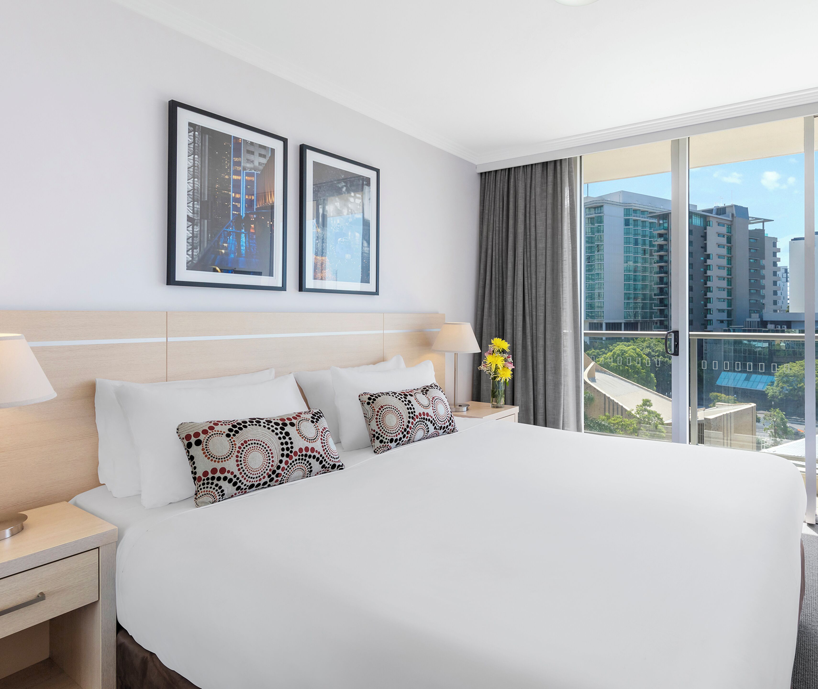 A hotel room with a large bed, two framed pictures on the wall, a nightstand with a lamp, and a view of buildings through the window.