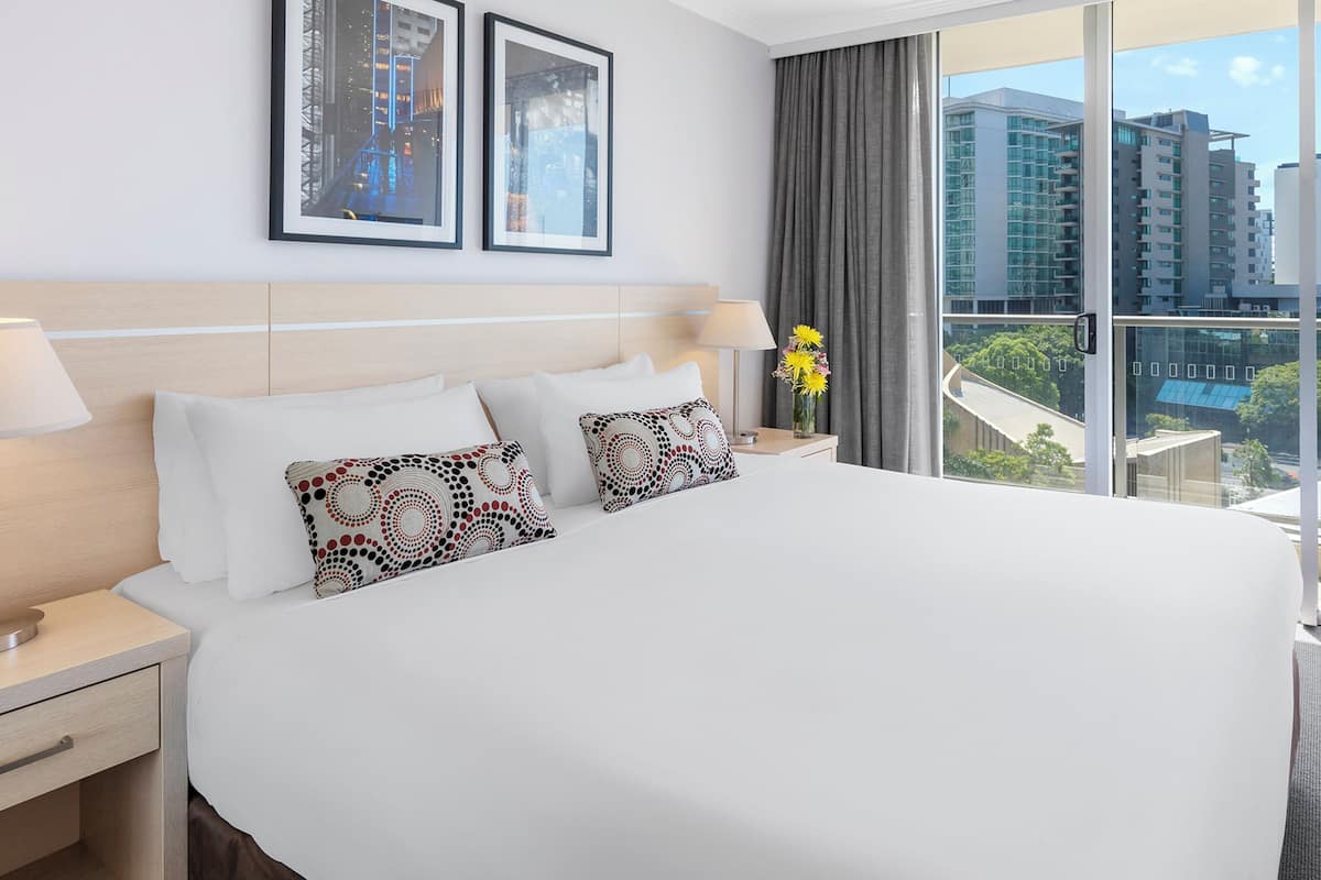 A hotel room with a large bed, two framed pictures on the wall, a nightstand with a lamp, and a view of buildings through the window.