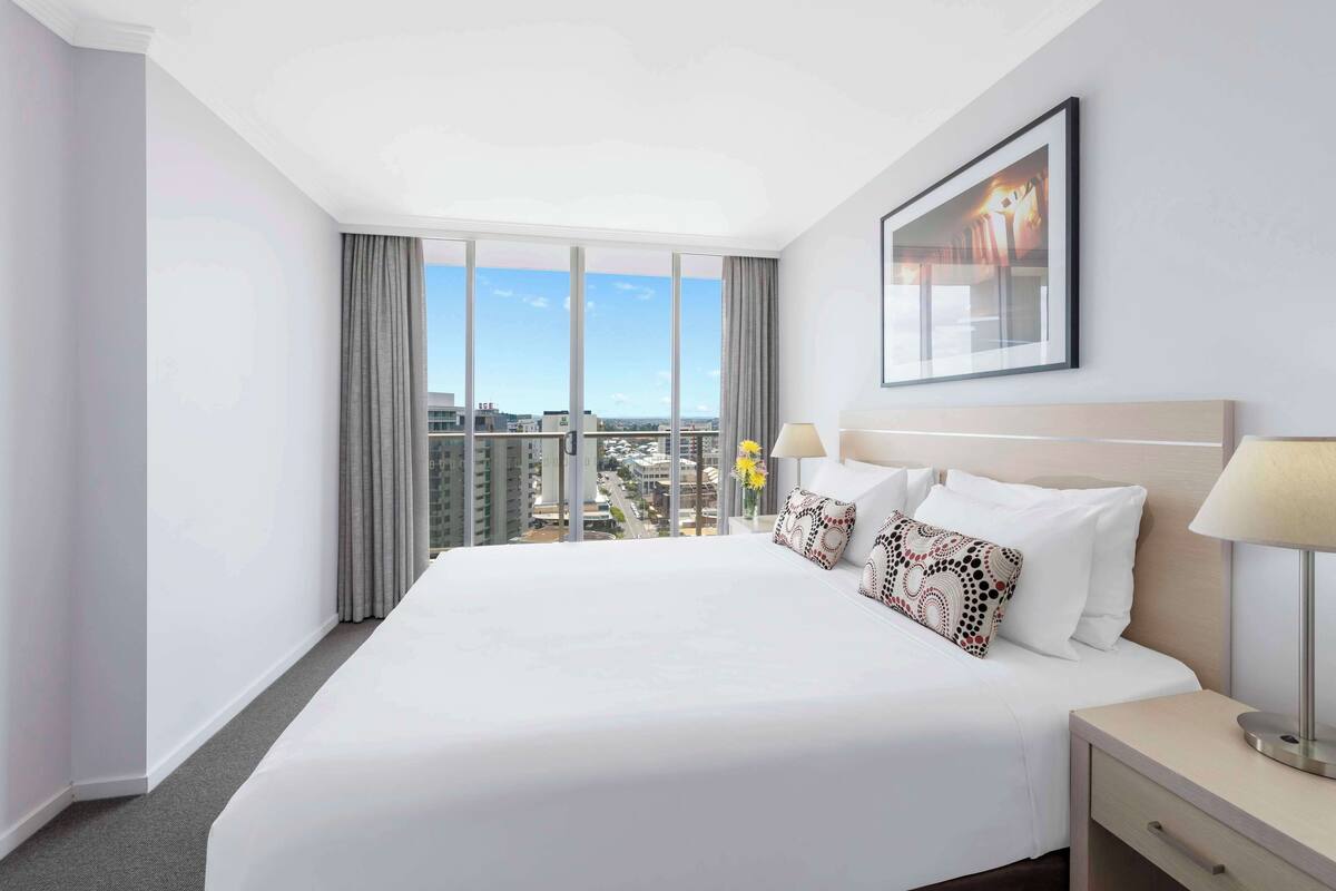 A hotel room with a large bed, a nightstand, a lamp, a framed picture, and a view of the city through large windows.