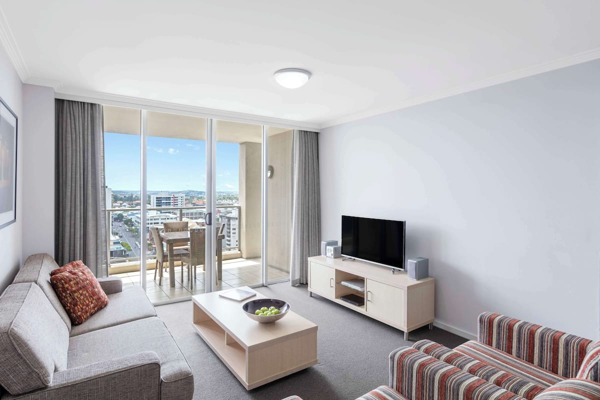 A modern living room with a sofa, a striped couch, a coffee table with a bowl, a TV on a stand, and a sliding glass door leading to a balcony with a city view.