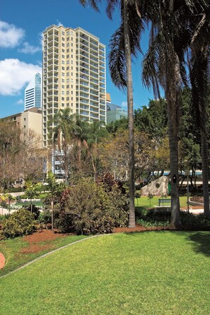 A park with a well-maintained lawn, palm trees, and a large building in the background.
