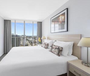 A hotel room with a large bed, two bedside lamps, a framed picture on the wall, and a view of the city through large windows.