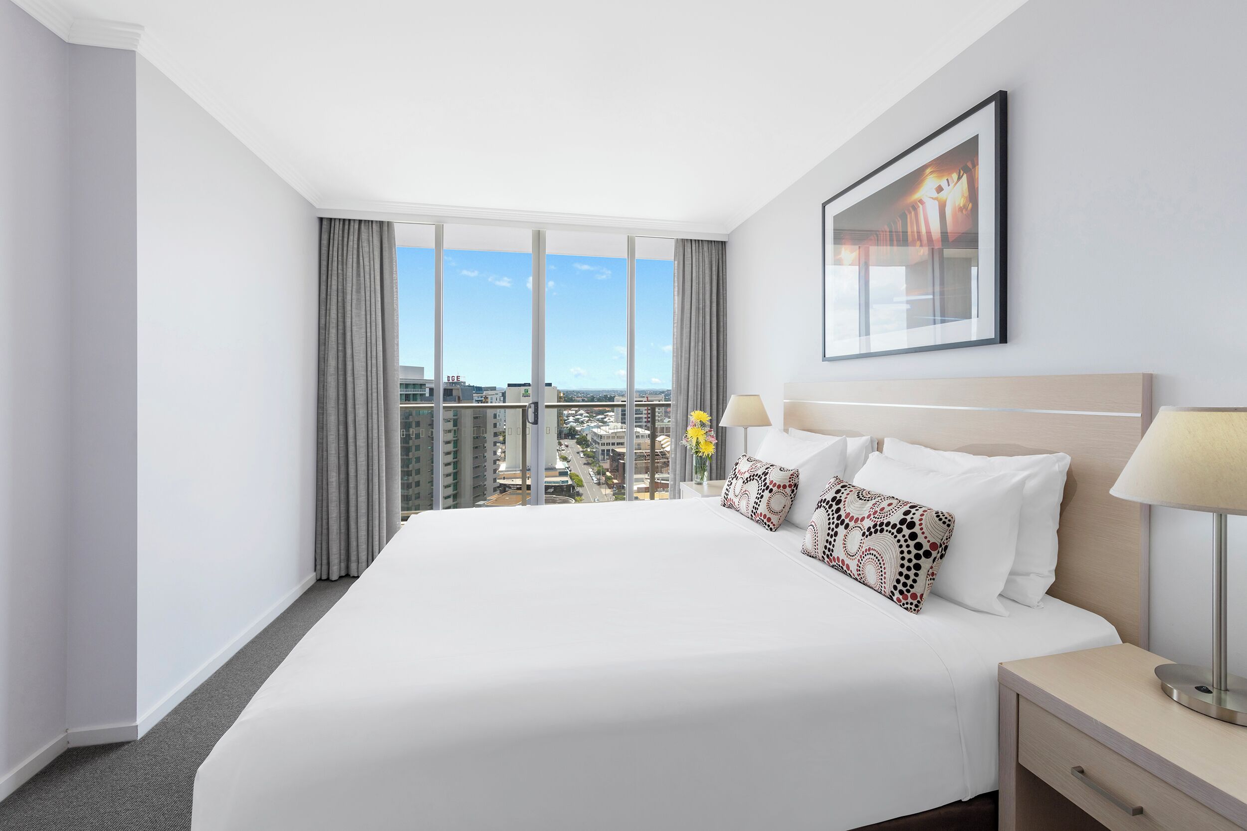 A hotel room with a large bed, a nightstand, a lamp, a framed picture on the wall, and a view of the city.