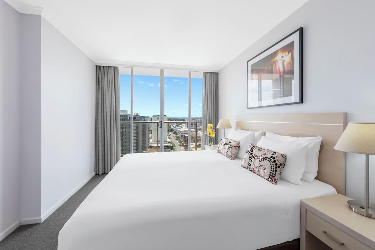 A hotel room with a large bed, a nightstand, a lamp, a framed picture on the wall, and a view of the city.