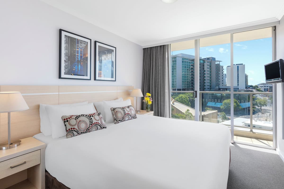 Habitación de hotel con una cama grande, dos cuadros en la pared, un televisor y un balcón con vista a la ciudad.