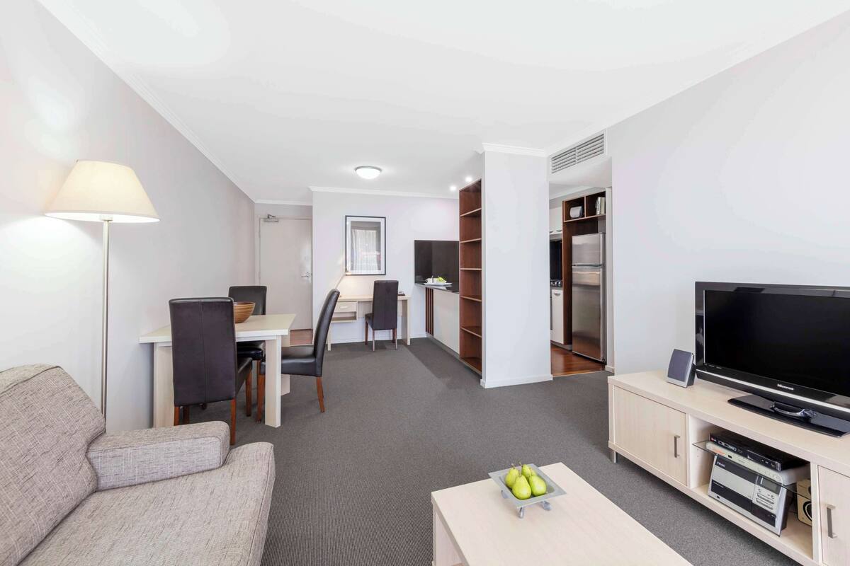 A modern hotel room with a sofa, a TV, a desk with a chair, a bookshelf, and a dining area.