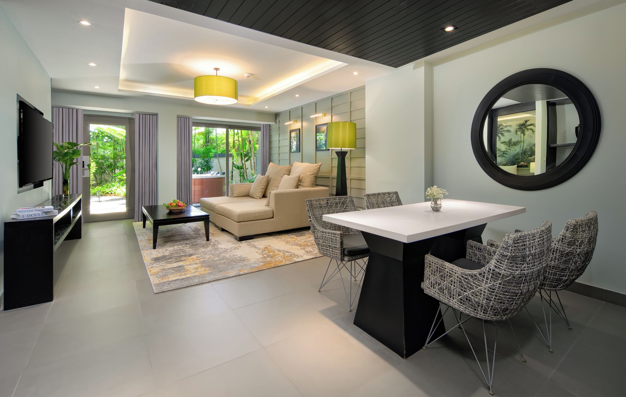 A modern living room with a sofa, coffee table, dining table, and chairs. There is a television on a stand, a lamp, and a large mirror on the wall.