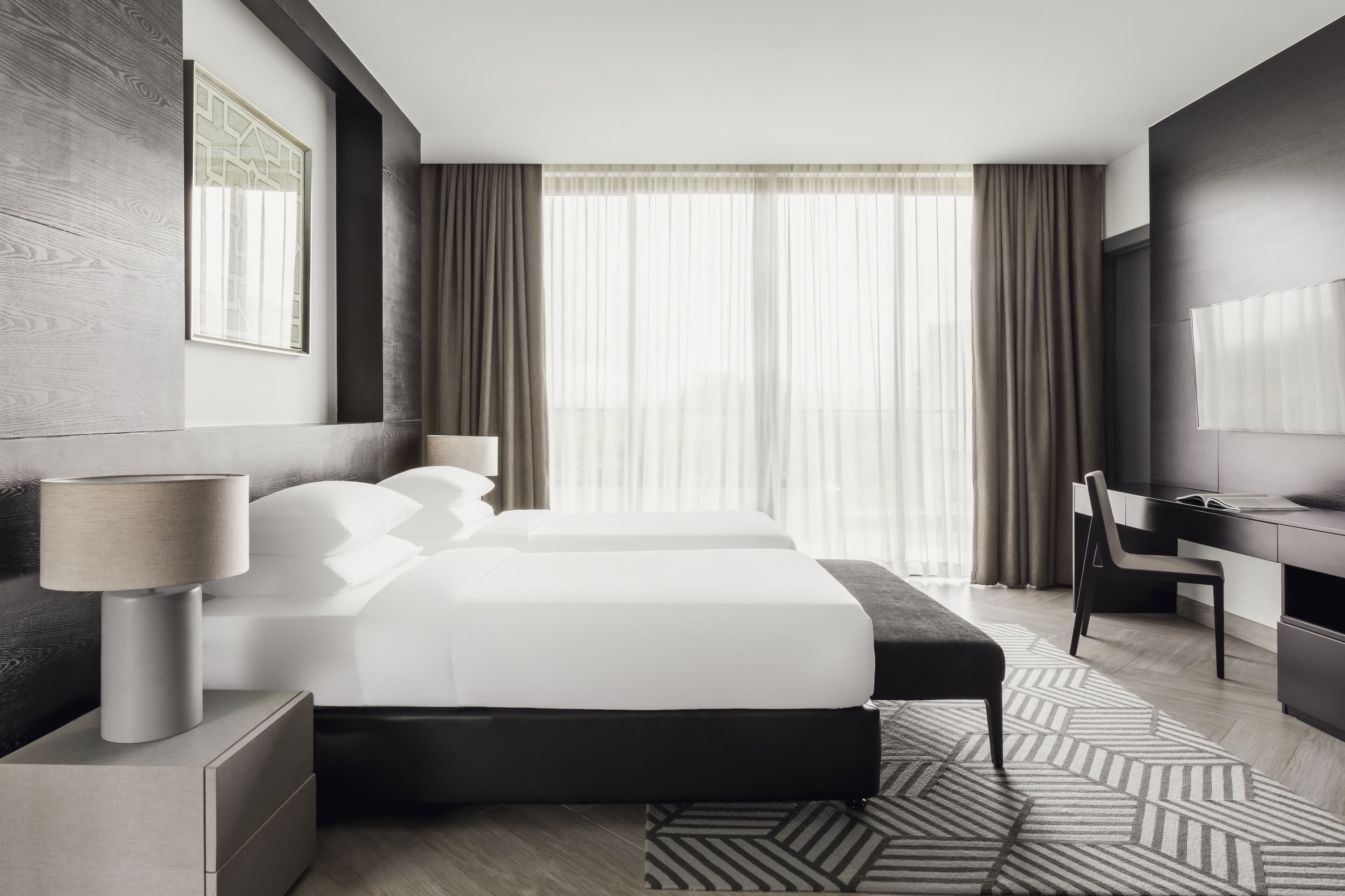 A modern hotel room with a large bed, bedside tables, a desk, and a view of the city through large windows.