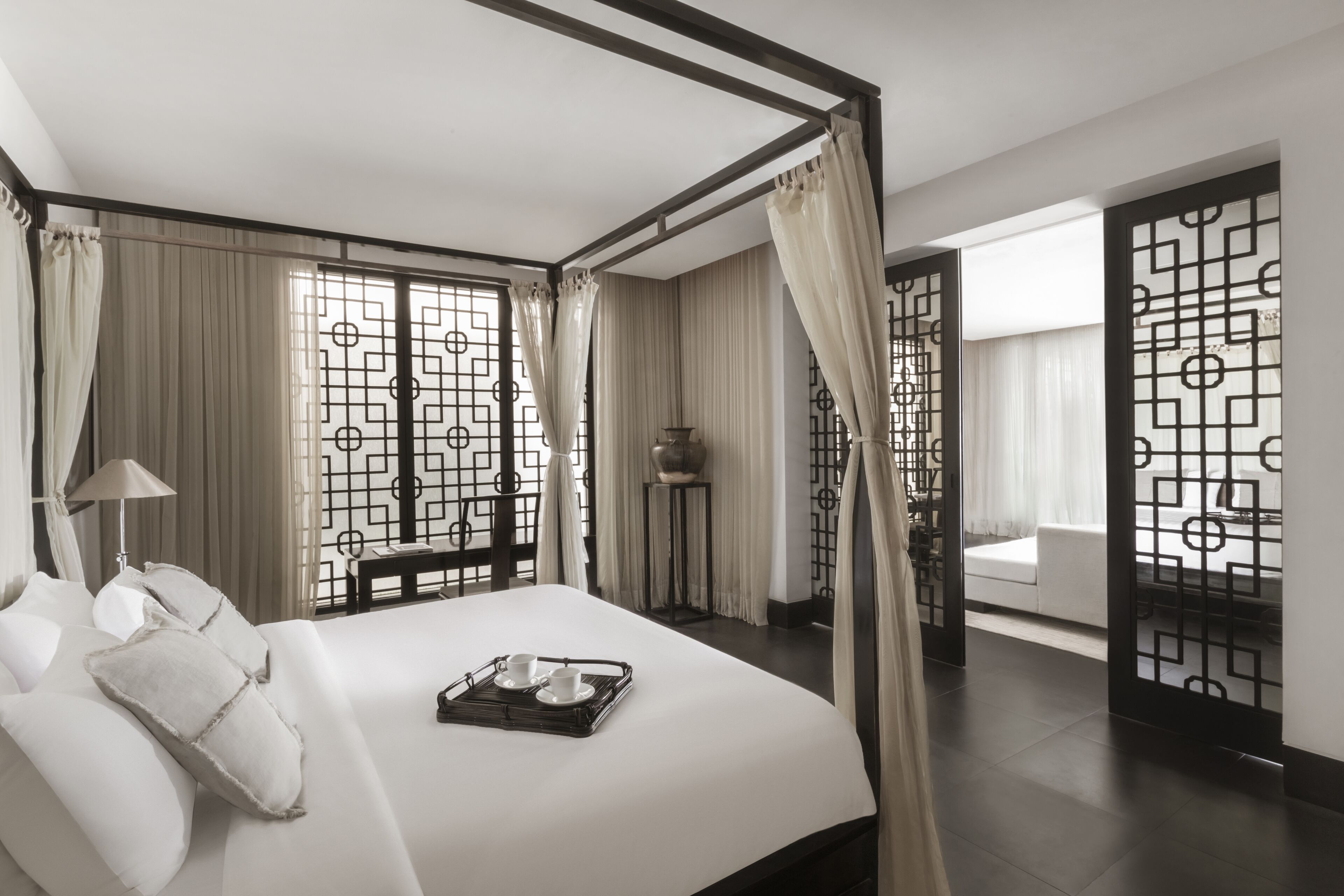 A modern hotel room with a four-poster bed, a tray with cups and saucers, and a view through a geometric-patterned screen to a balcony.