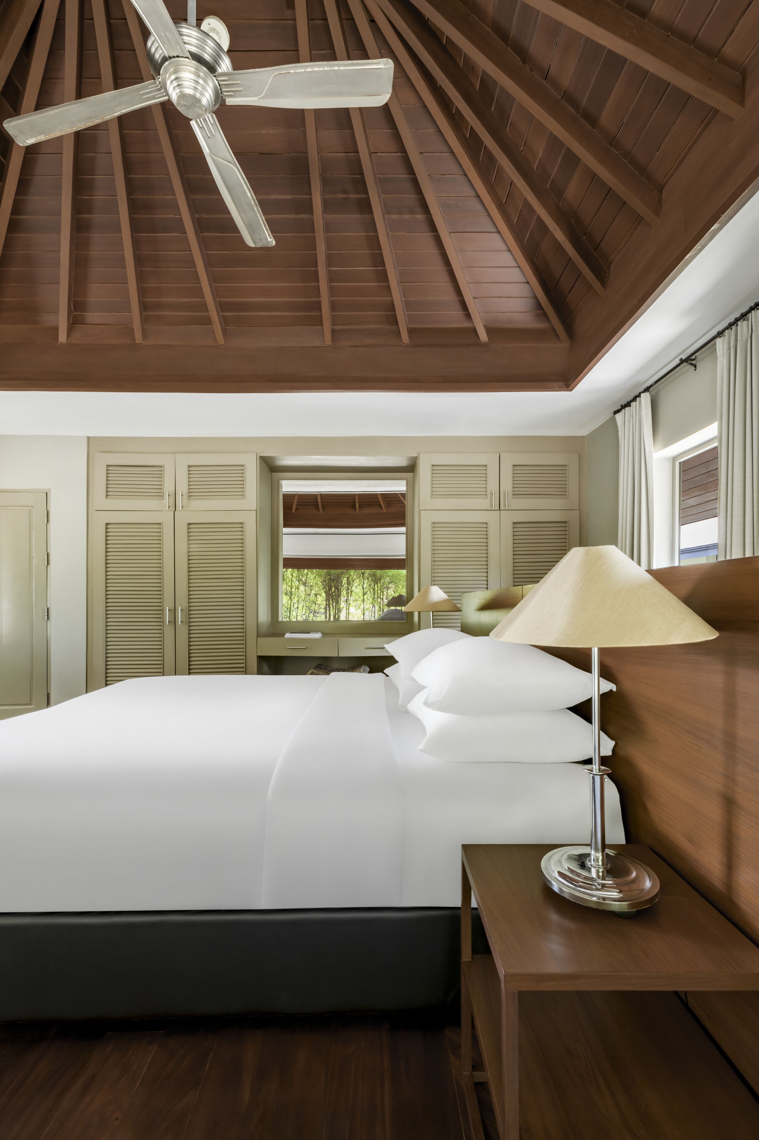 Une chambre avec un plafond en bois, un lit recouvert de draps blancs, une table de chevet avec une lampe et une fenêtre avec des volets.