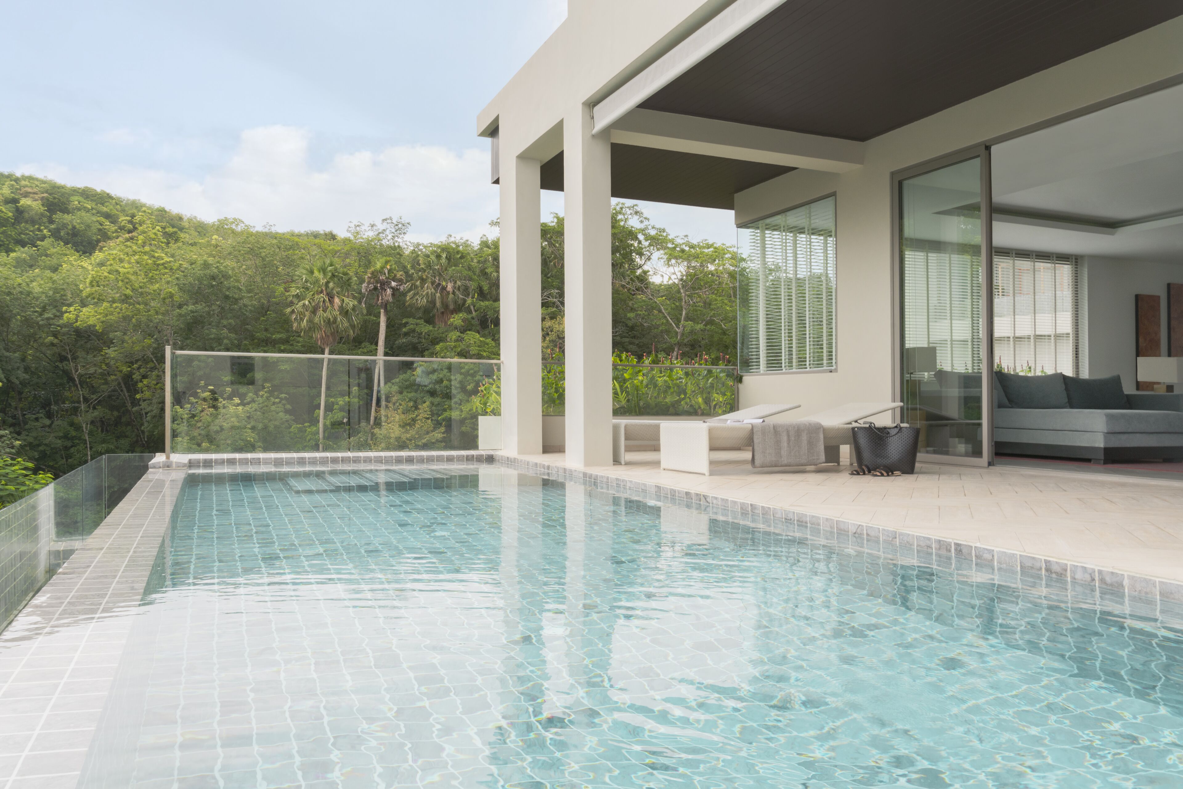 A modern outdoor swimming pool with clear blue water, surrounded by a glass fence and a white tiled deck.