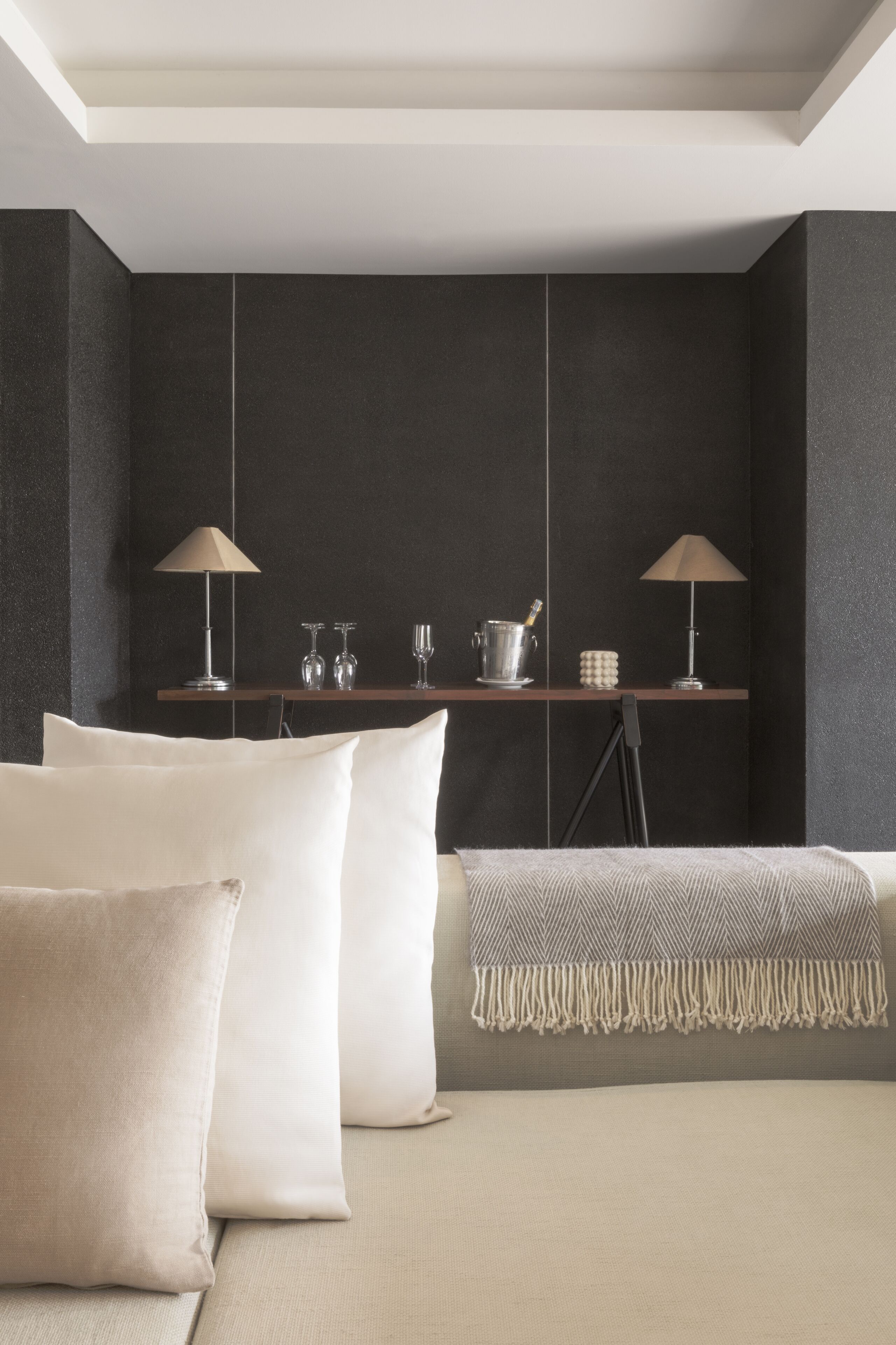 A modern bedroom with a neutral color palette, featuring a bed with white linens, two bedside lamps, a small shelf with decorative items, and a dark accent wall.