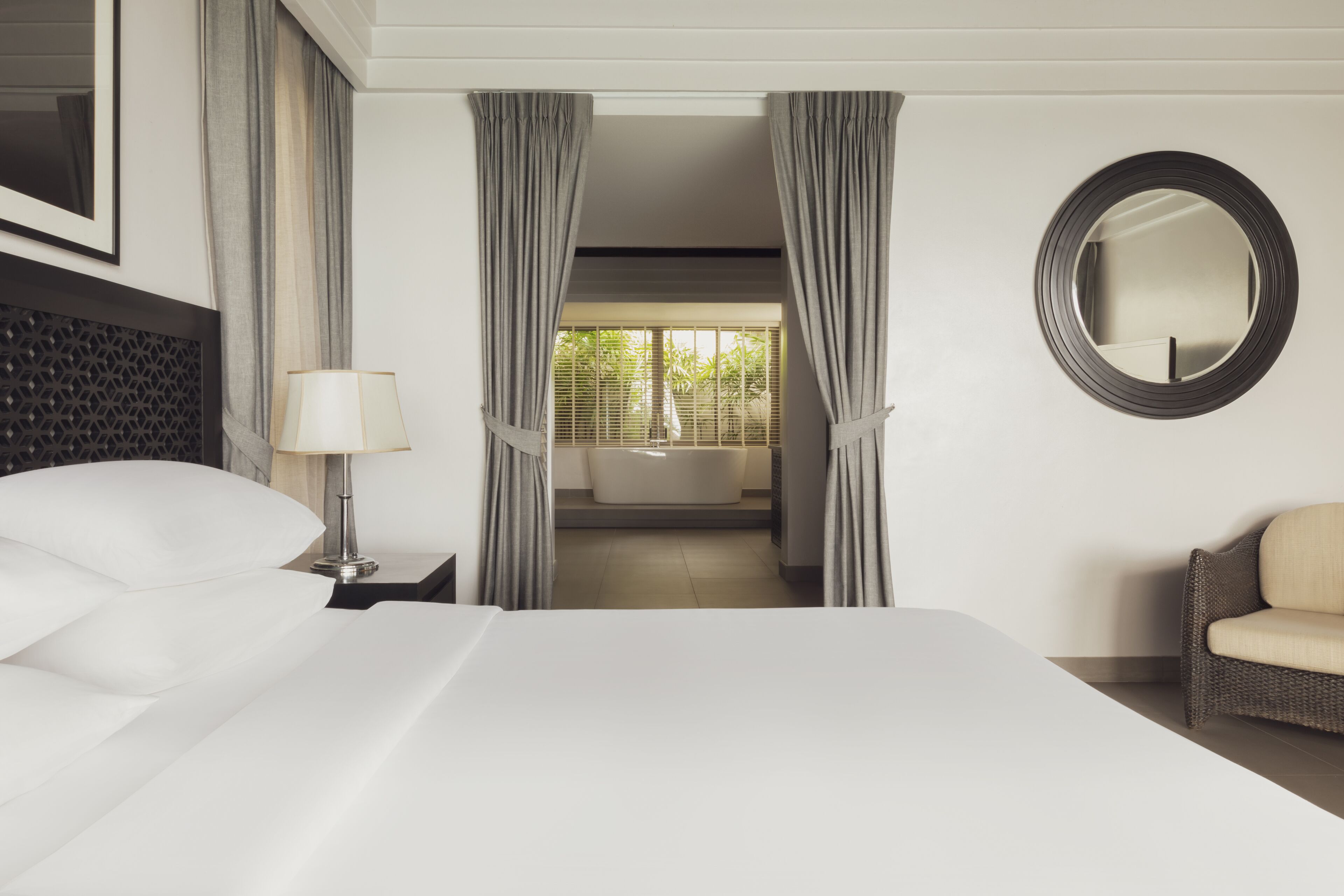 Une chambre d’hôtel avec un grand lit, une table de chevet avec une lampe, une chaise, un miroir rond et une vue sur une salle de bain avec une baignoire.