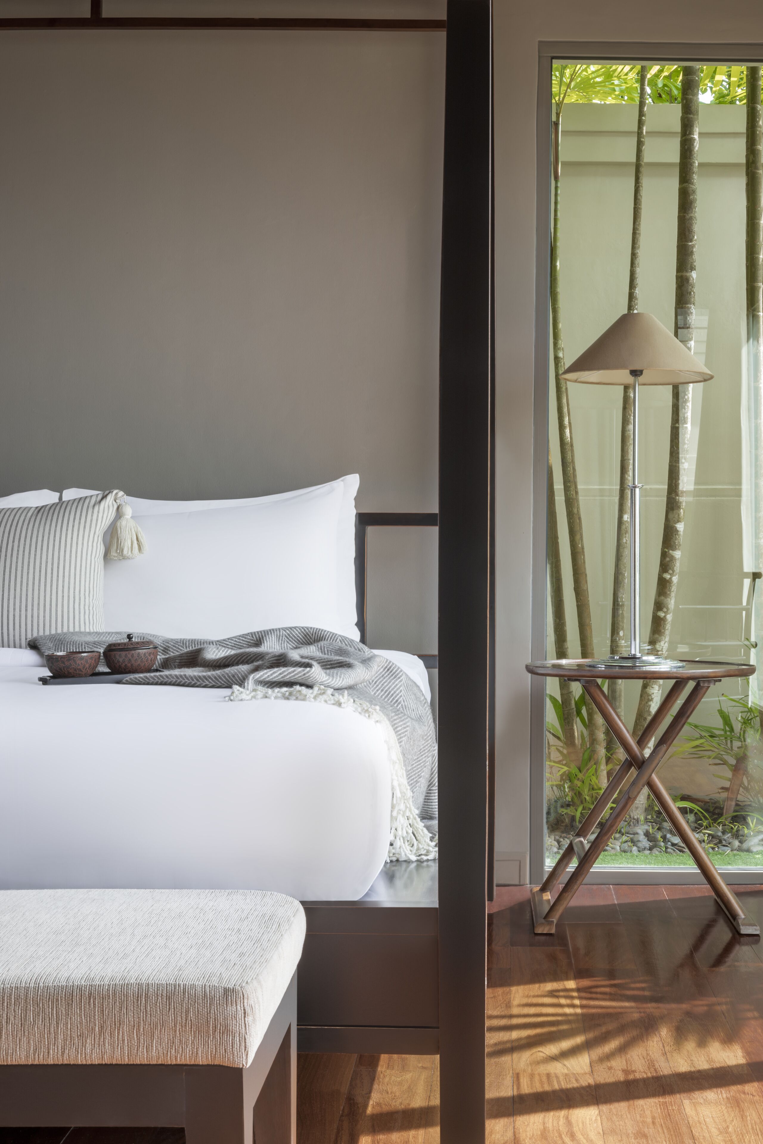 Une chambre à coucher avec un lit, une table de chevet, une lampe et une vue sur des bambous, visible à travers une porte vitrée.
