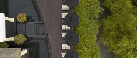 An aerial view of a rooftop terrace with a row of white lounge chairs and black umbrellas, surrounded by greenery and a paved walkway.