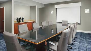 Un salón de conferencias con una mesa de vidrio, sillas grises, un mueble de madera con máquinas de café y una pantalla para proyector.