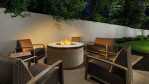 A circular fire pit surrounded by wicker chairs and a table, set against a backdrop of a white wall and lush greenery. - Courtyard By Marriott Farmington (Newington)