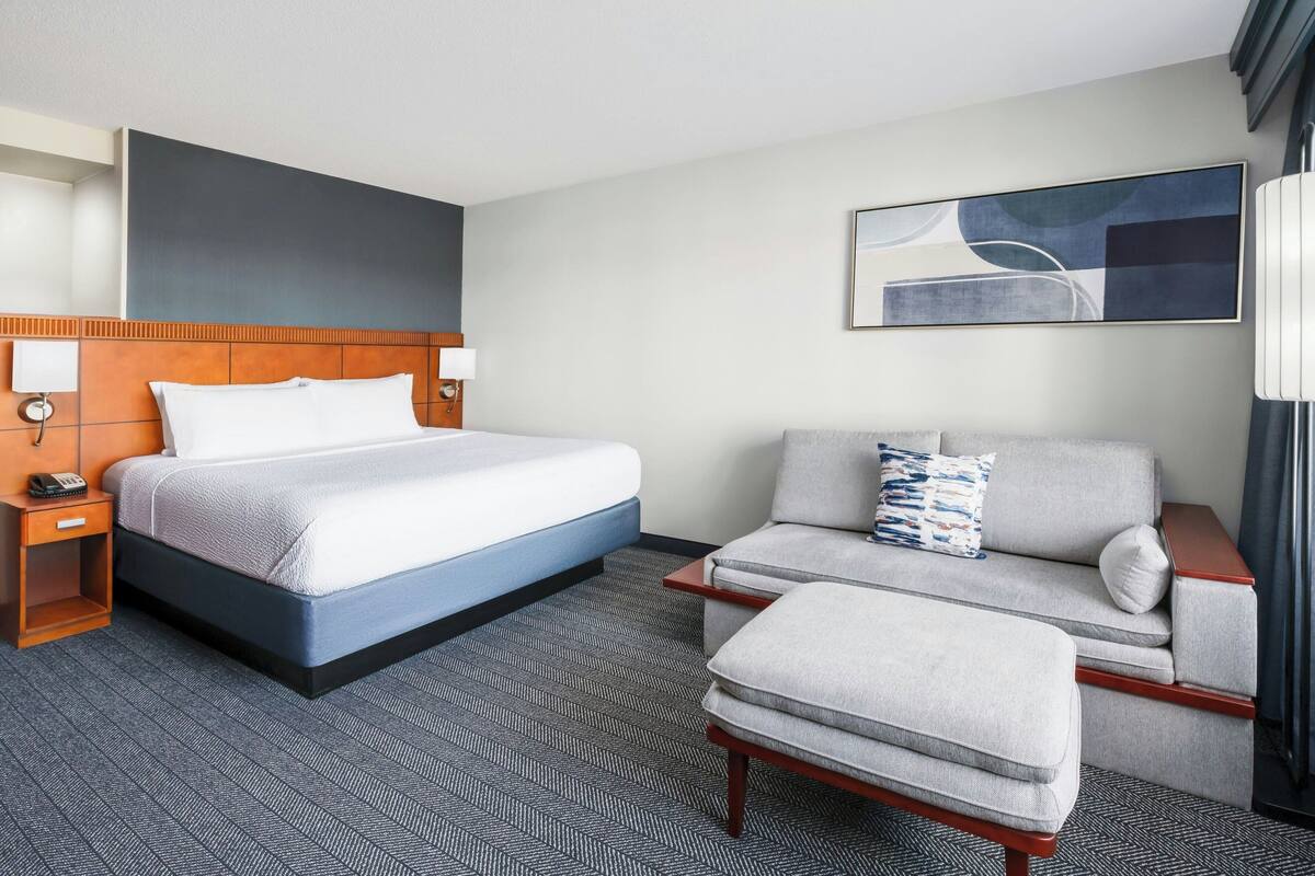 A hotel room with a bed, a sofa, an ottoman, a nightstand, and a wall-mounted artwork.