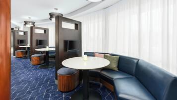 A modern hotel room with a blue patterned carpet, a round white table, a blue leather sofa, and a TV mounted on a dark panel.
