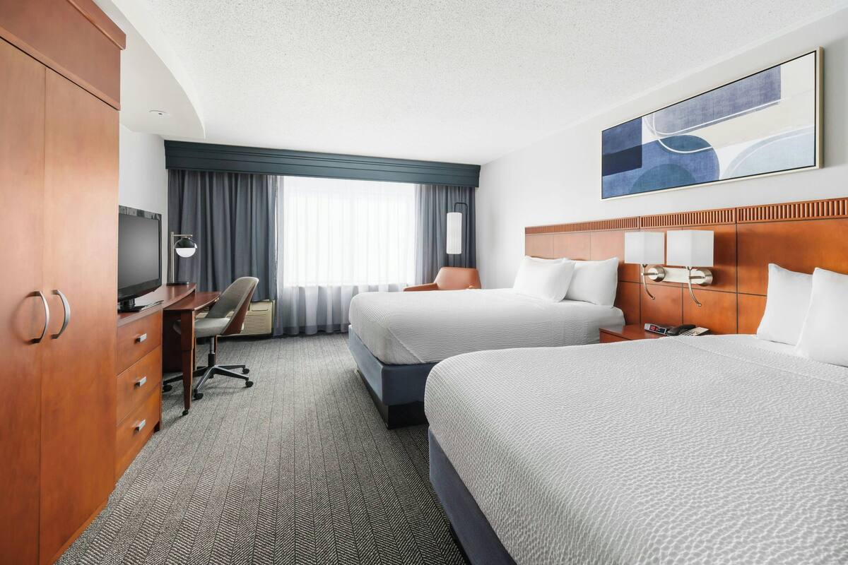 A hotel room with two beds, a desk with a chair, a TV, and a large window with curtains.