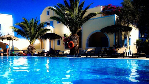 A swimming pool with lounge chairs and palm trees, surrounded by buildings with arched windows and balconies. - Hotel Zephyros (Santorini)