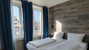 Habitación de hotel con dos camas, cabecera de madera, vistas de edificios y una ventana con cortinas azules.