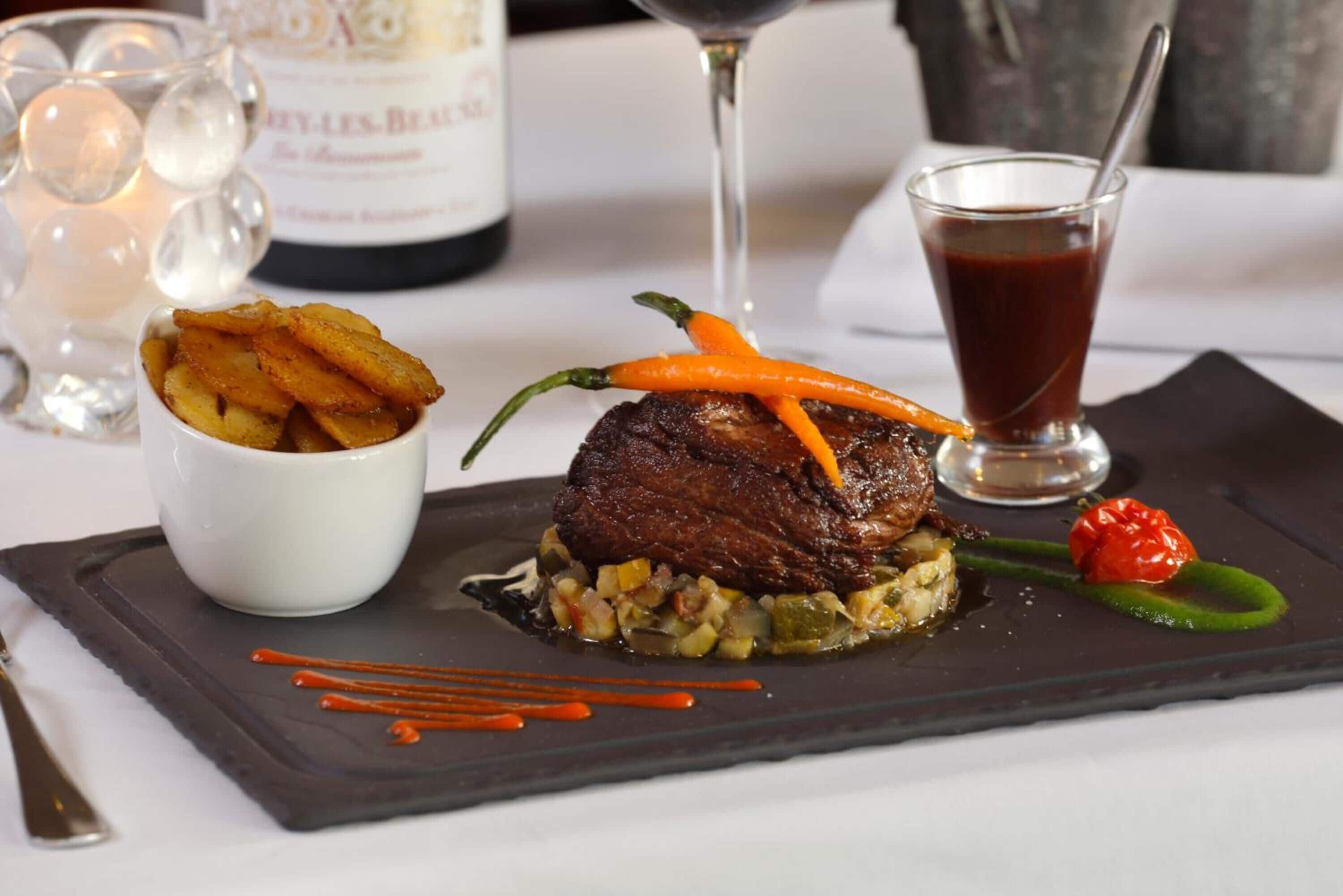 Une assiette garnie d’un steak, de légumes et d’une petite portion de frites, accompagnée d’un verre de vin rouge et d’une bouteille en arrière-plan.