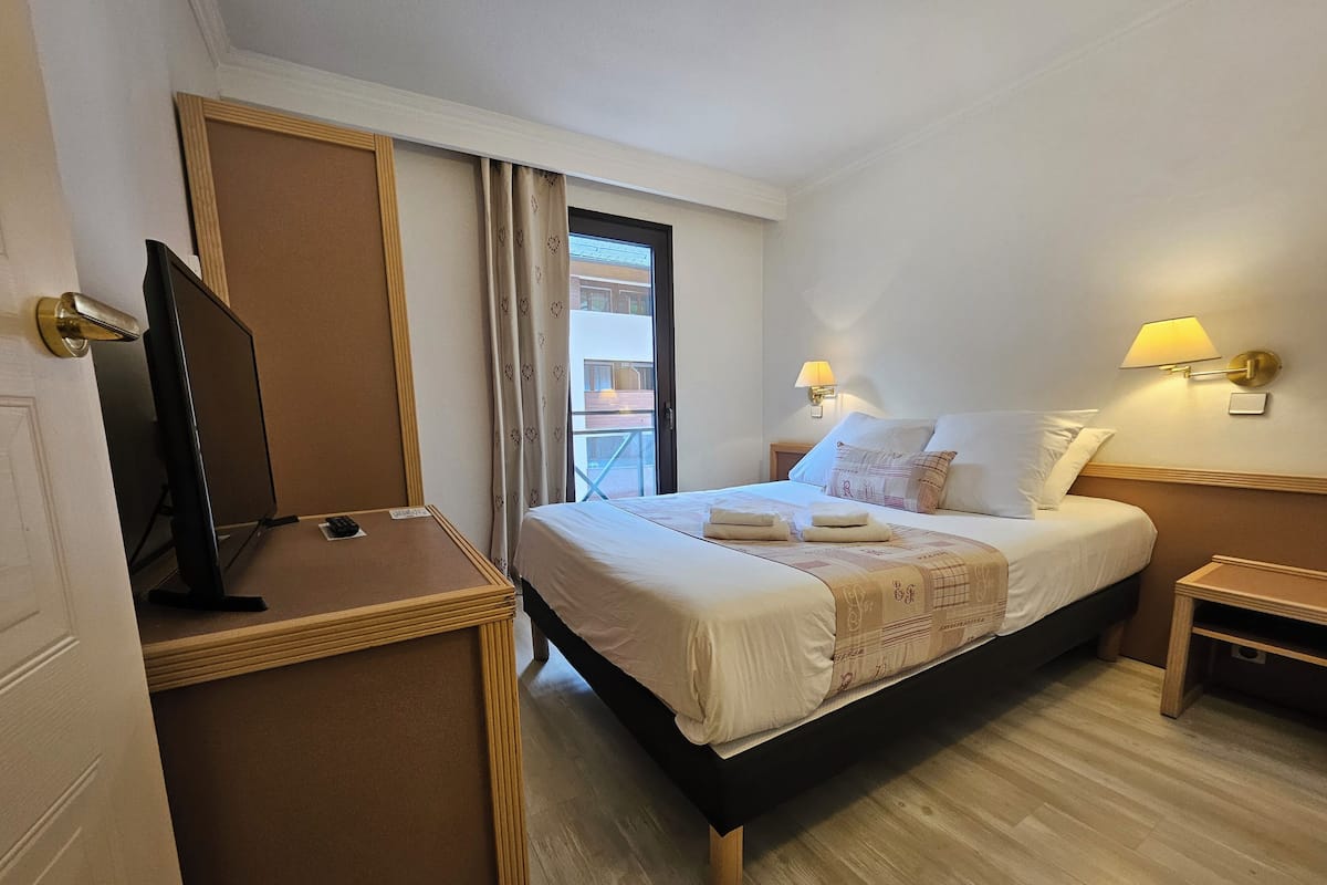 Una habitación de hotel con cama, lámparas de noche, televisor sobre un mueble y ventana con cortinas.