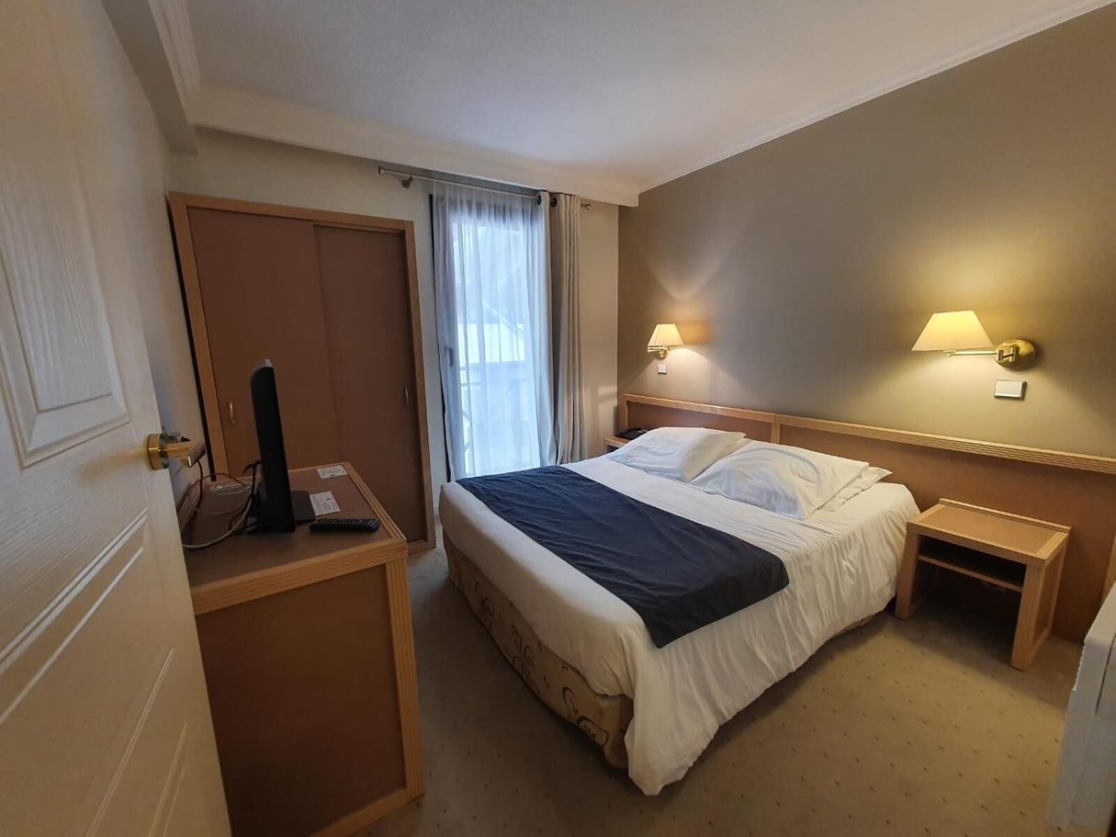 A hotel room with a bed, bedside tables, a wardrobe, and a window with curtains.
