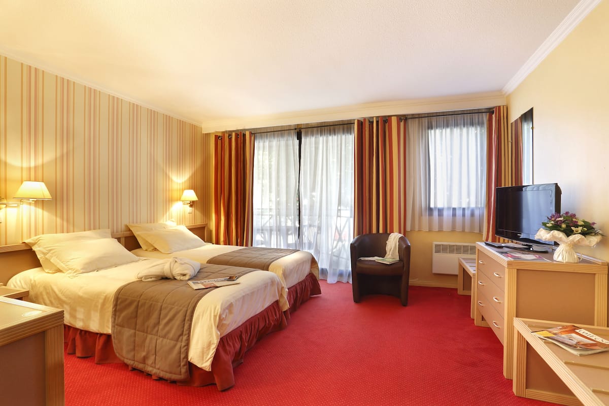 Habitación de hotel con dos camas, un escritorio con televisor, una silla y una pequeña mesa con un florero.
