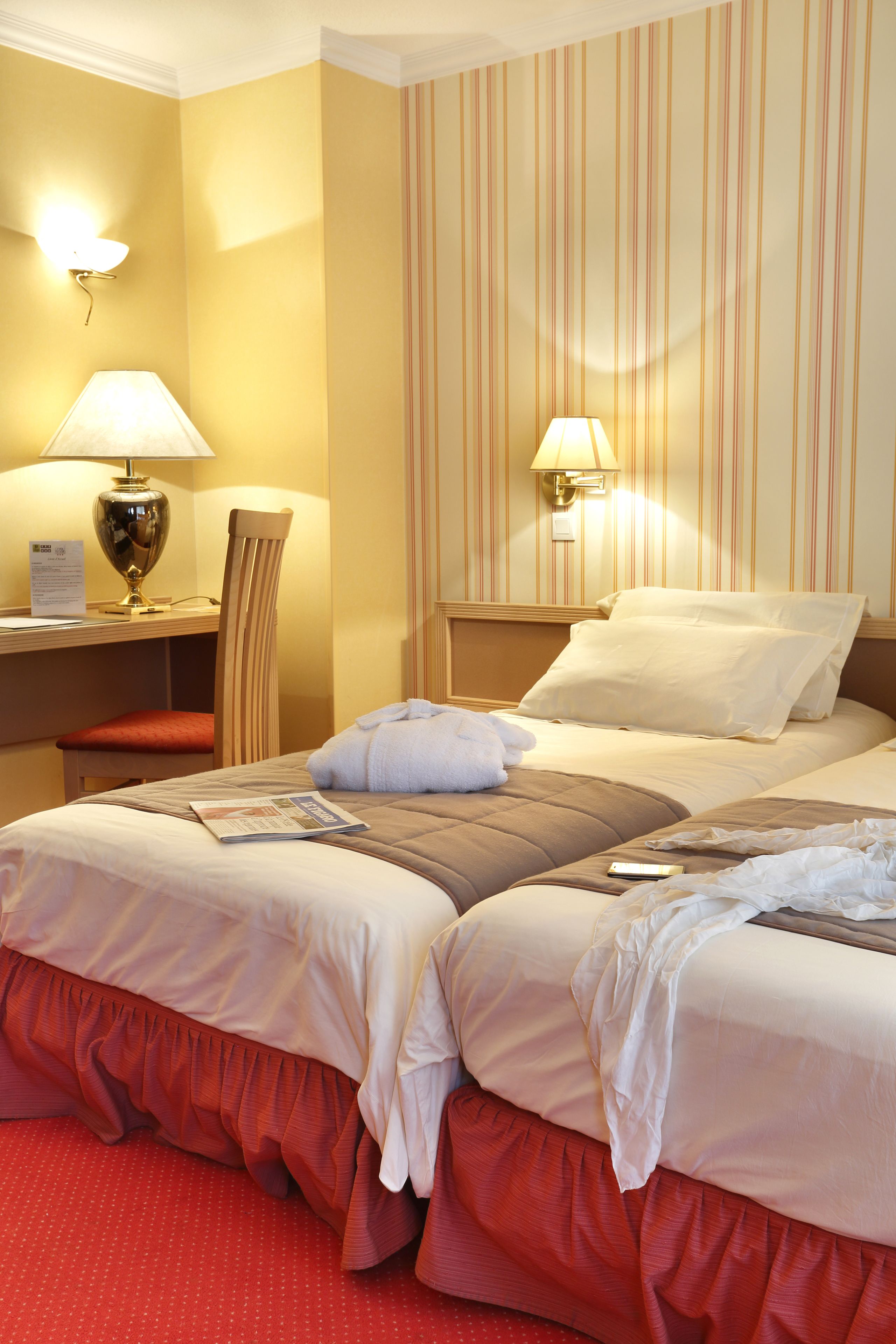 Ein Hotelzimmer mit zwei Betten, einem Schreibtisch mit Lampe, einem Sessel und gestreifter Tapete.