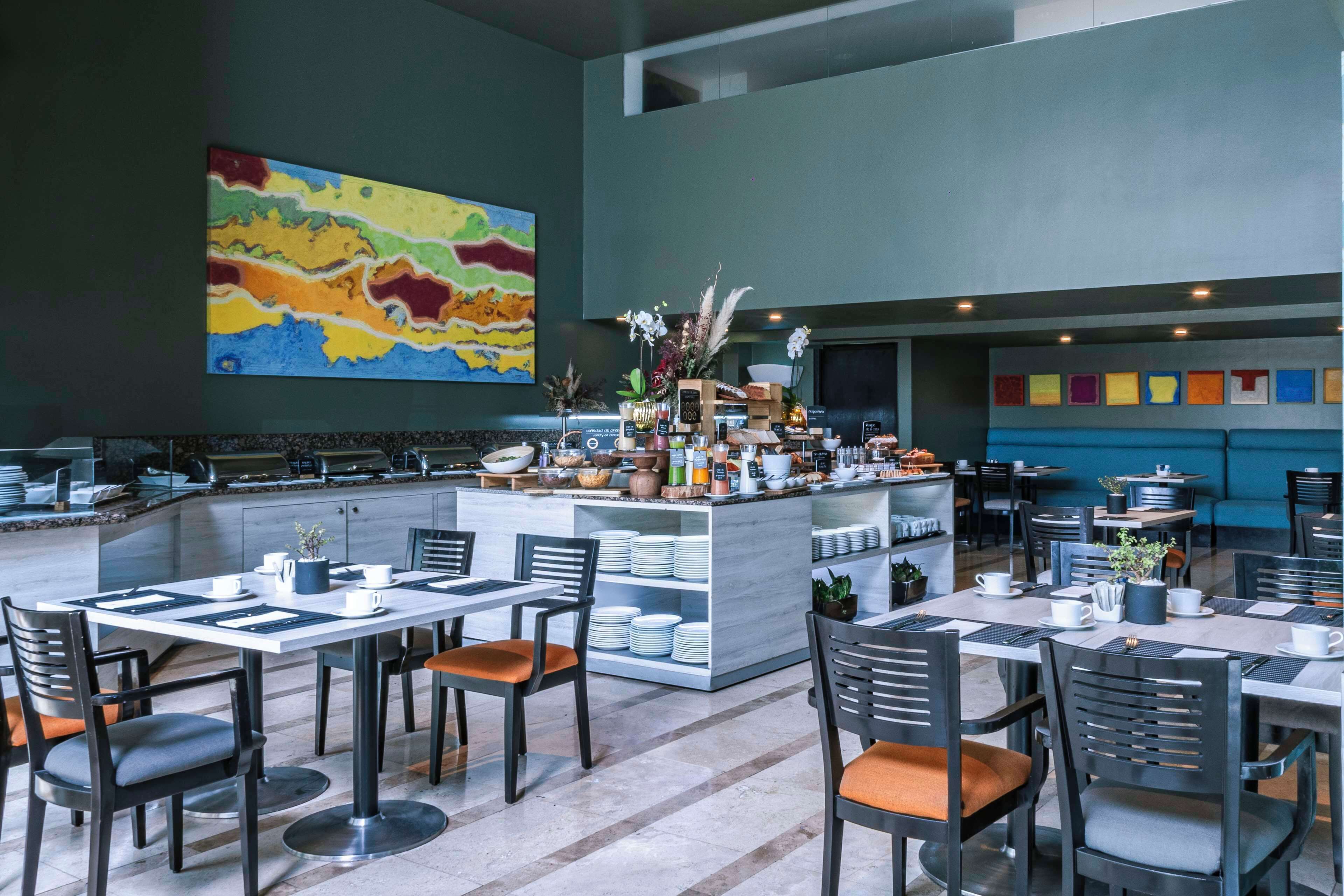 Un restaurante moderno con una barra central, varias mesas para comer y sillas. Hay un cuadro abstracto y colorido en la pared, y una variedad de plantas y flores en la barra.